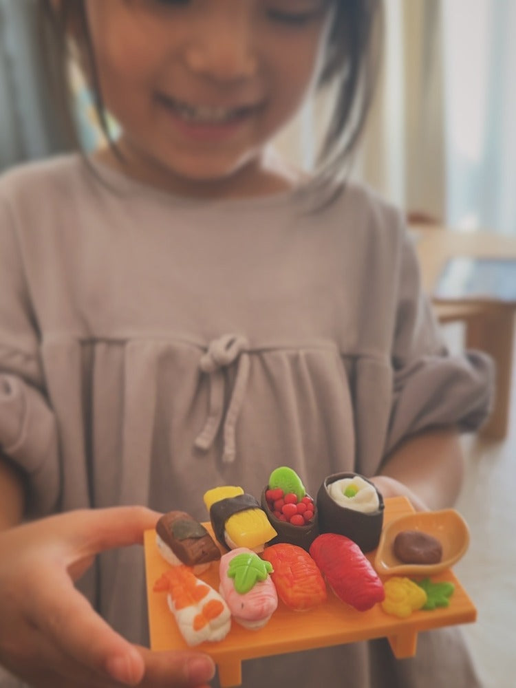 粘土で作った色とりどりのお寿司を、黄色い台に並べて手に持つ子ども。