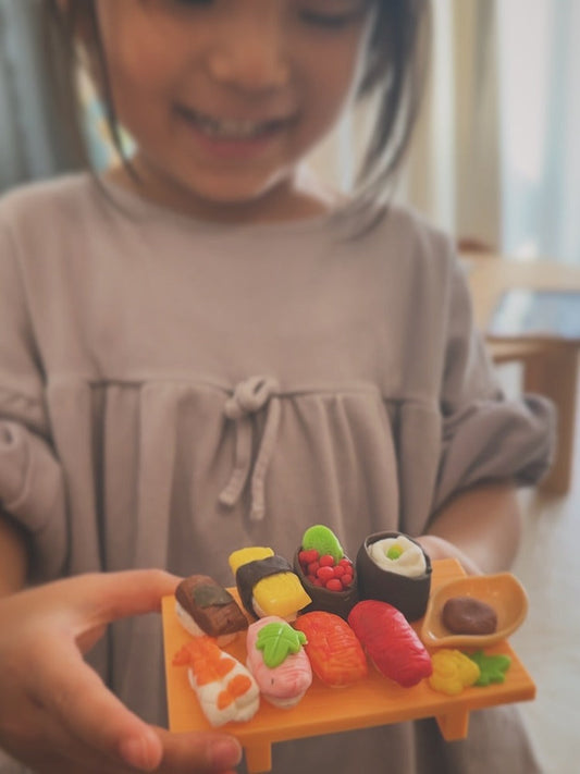 粘土で作った色とりどりのお寿司を、黄色い台に並べて手に持つ子ども。